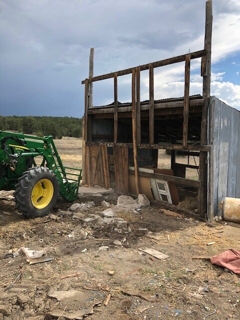 Chicken coop demolition — John Deere tractor with front loader, salvaged boards piled in foreground, Mora River valley beyond