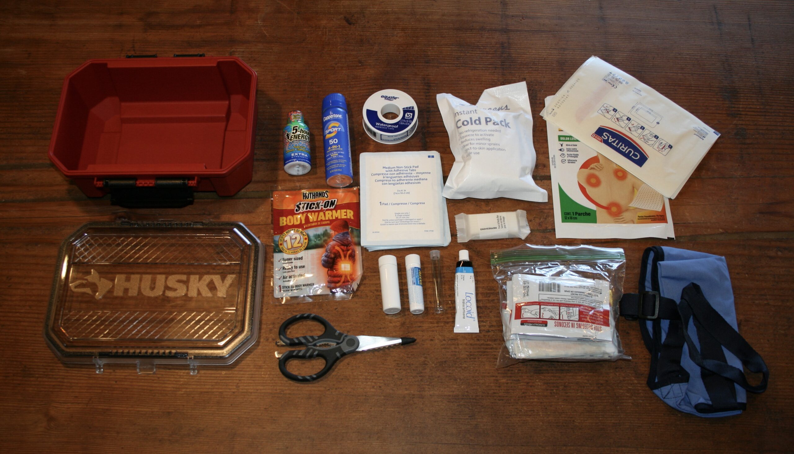 Husky hard case open with first aid contents spread including trauma scissors, cold pack, gauze, tourniquet, mylar blanket, and Locoid